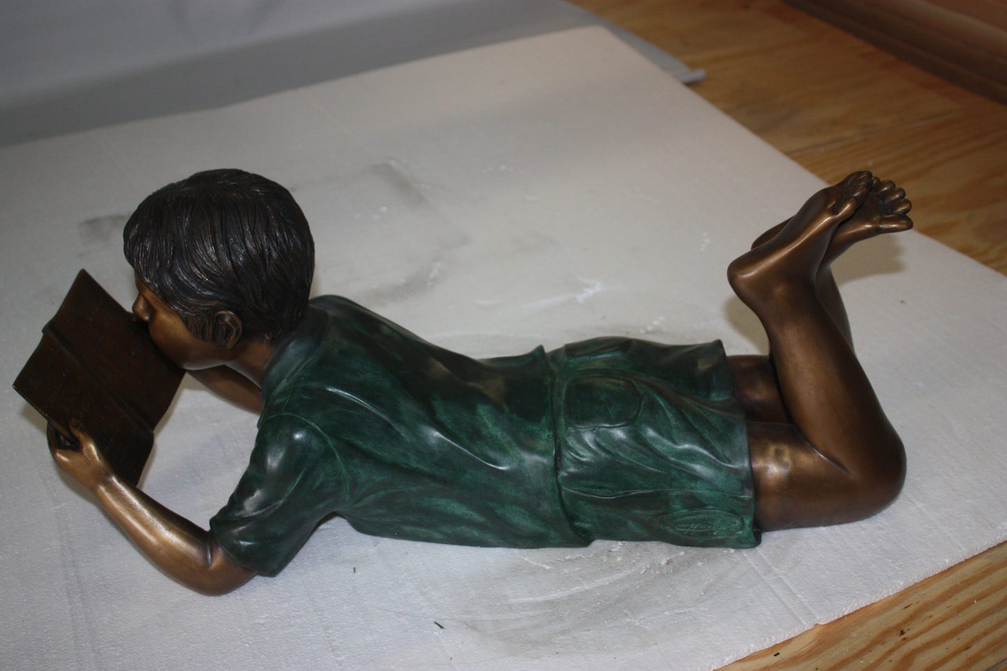 boy laying reading book bronze Statue - Size: 24"L x 10"W x 11"H.